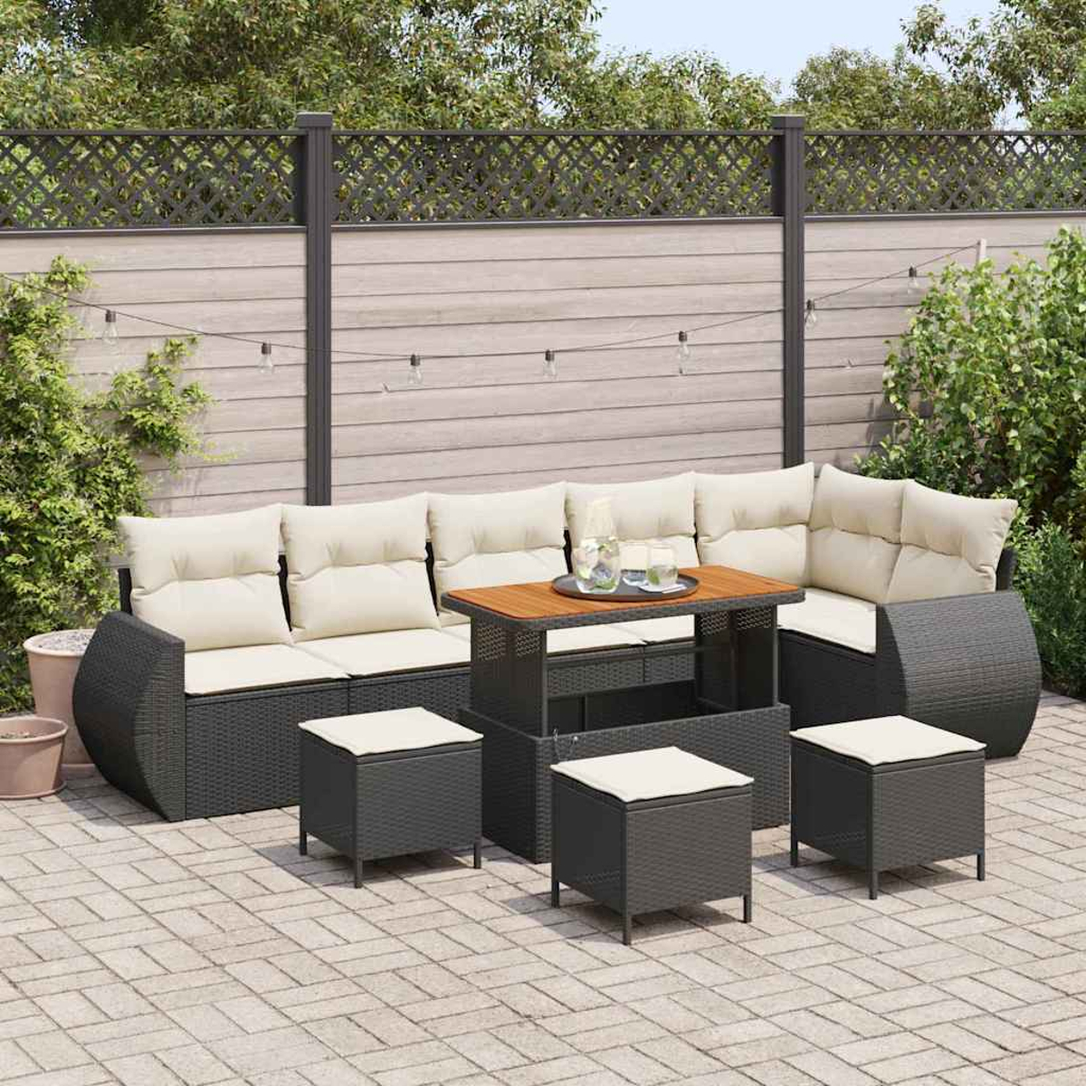 Set di mobili da giardino. Divano in vimini grigio con cuscini color crema, tavolo in legno e sgabelli piccoli. Una recinzione in legno e piante sullo sfondo.