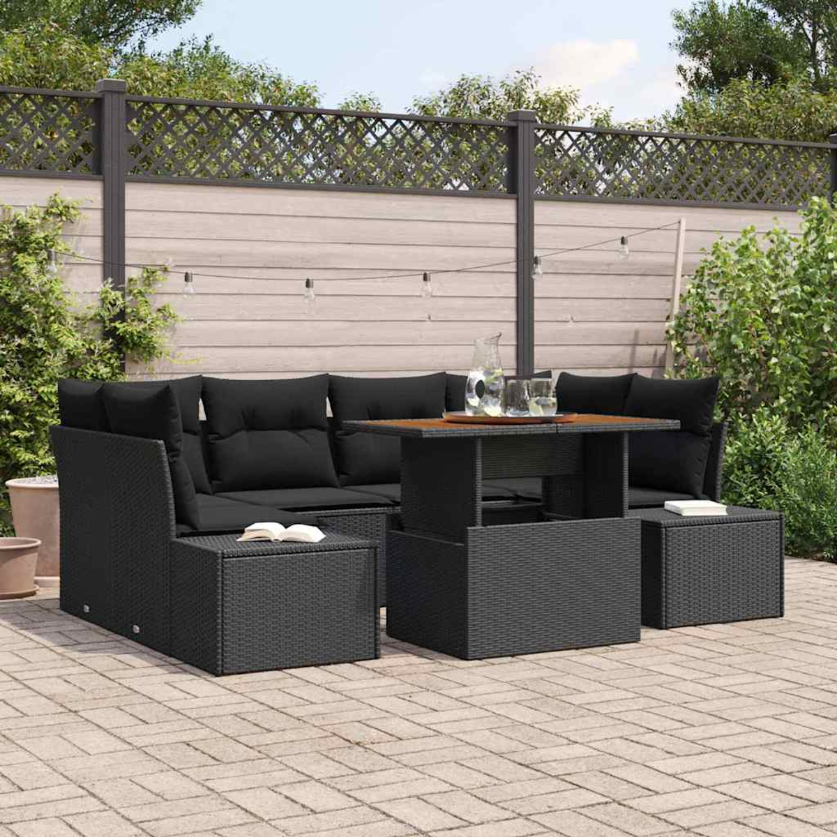 Terrassengarnitur im Freien mit schwarzen Kissen und Tisch. Hintergrund mit Holzzaun, Grün und Lichterketten.