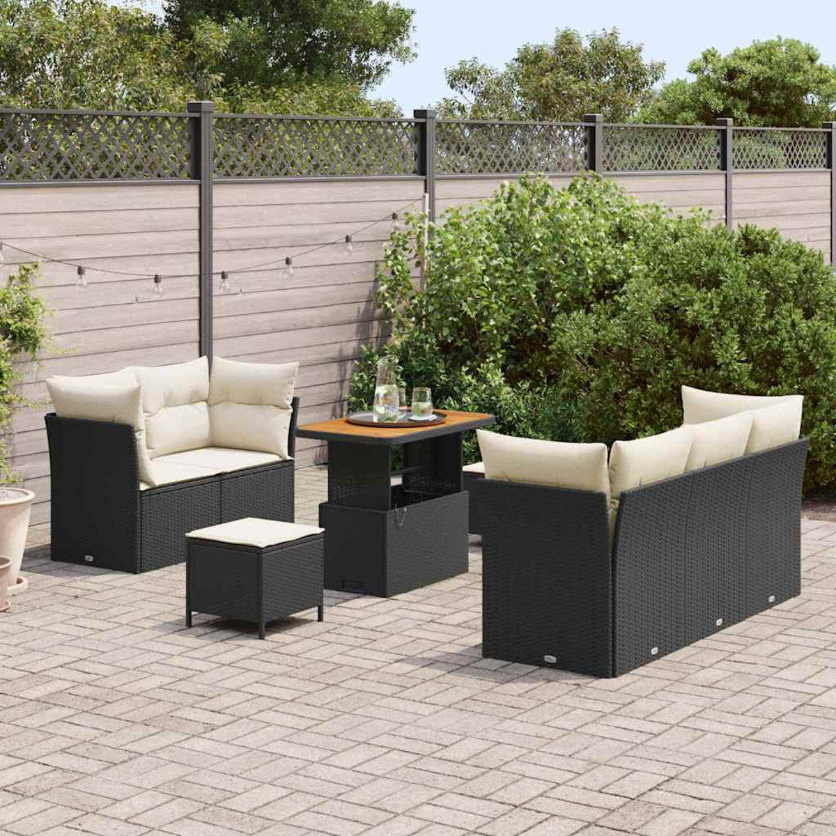 Terrassengarnitur mit schwarzen Korbmöbeln und beigen Kissen, einschließlich Tisch und Zaun mit Grün im Hintergrund.