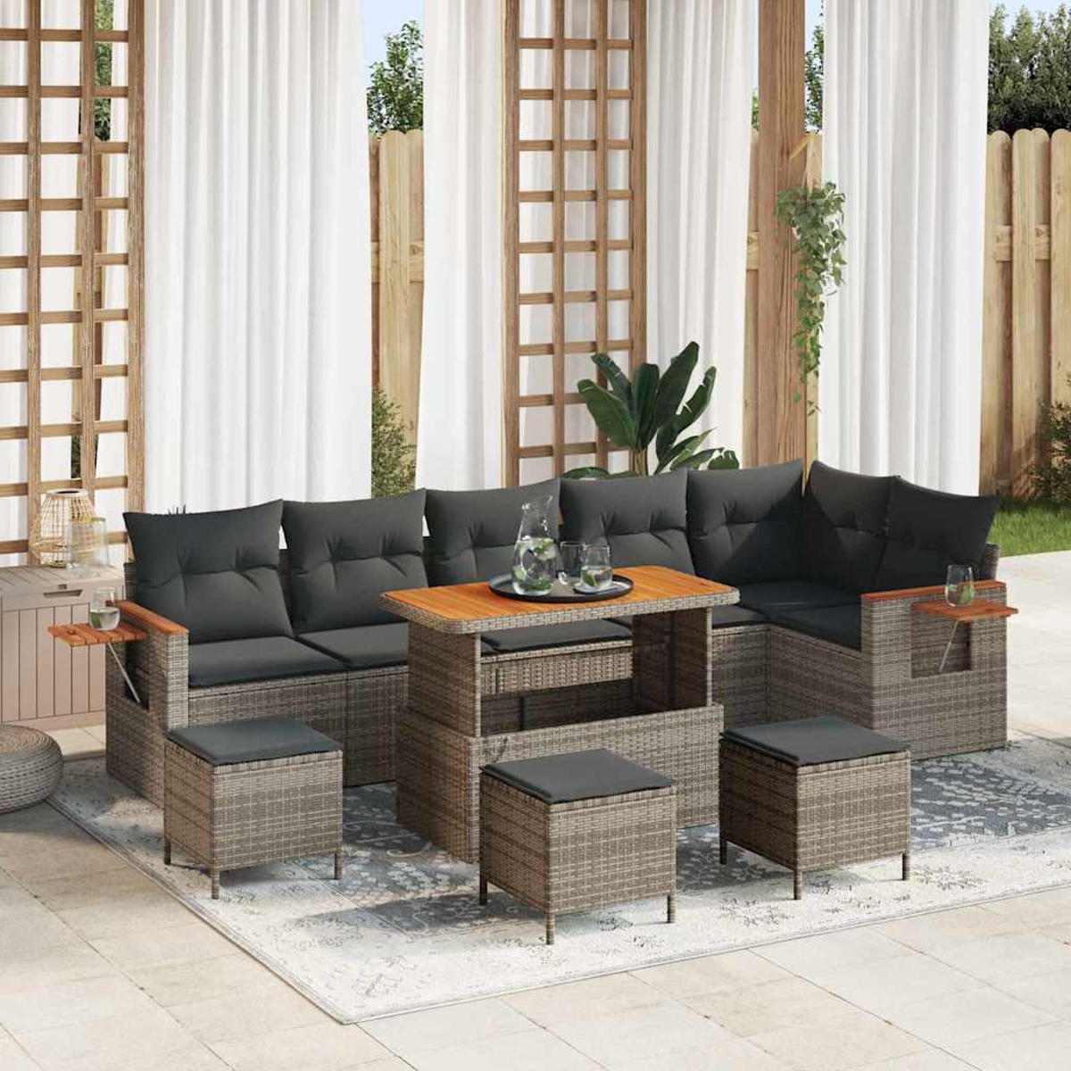 Set di mobili da esterno: divano, pouf e tavolo. Cuscini grigi e telai intrecciati. Tende bianche sullo sfondo. Accenti in legno.