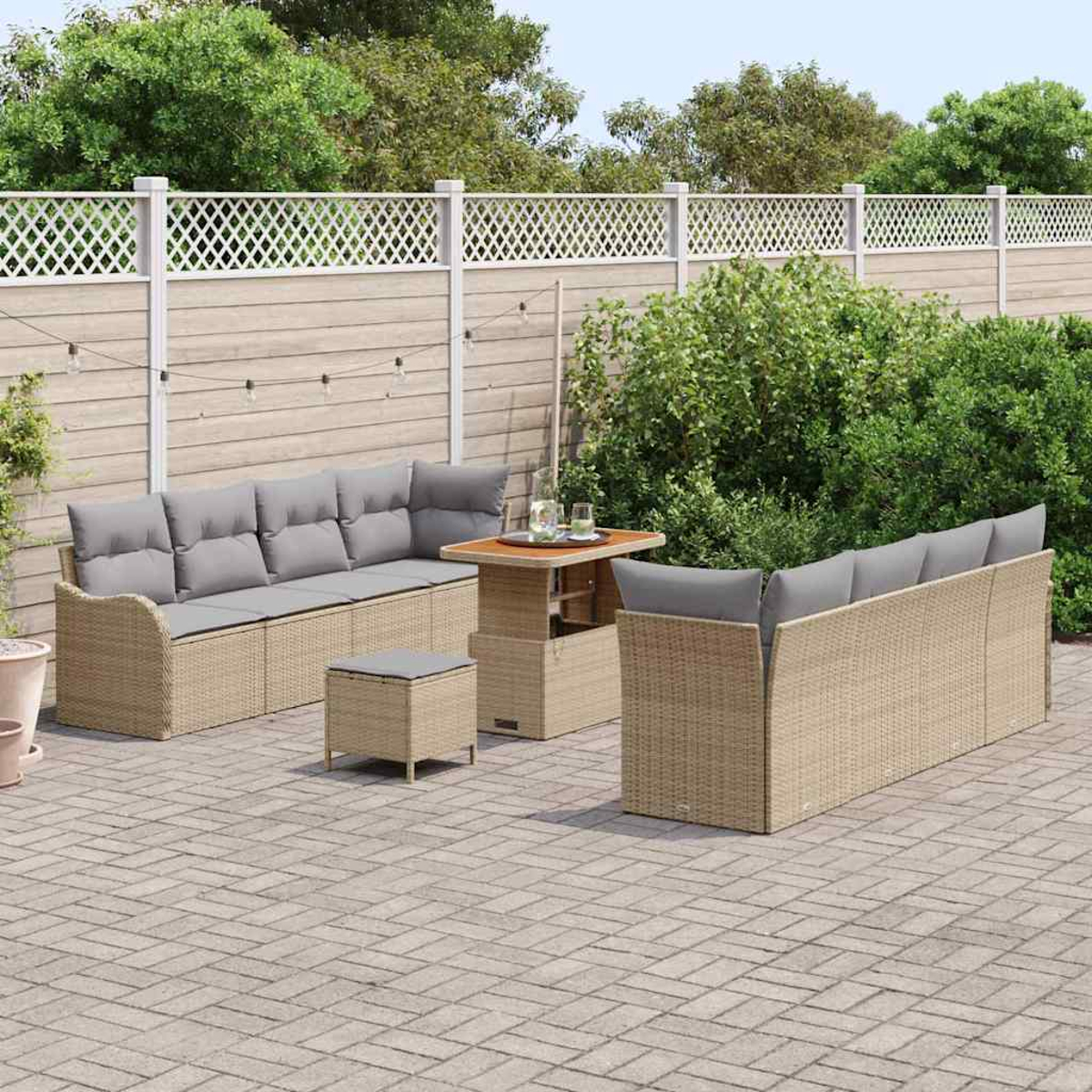 Terrassenset mit Korbs Sofas, Ottomane und Tisch. Graue Kissen, tanfarbenes Korbgeflecht, auf einer Ziegelstein-Terrasse, mit weißem Zaun und Grün.