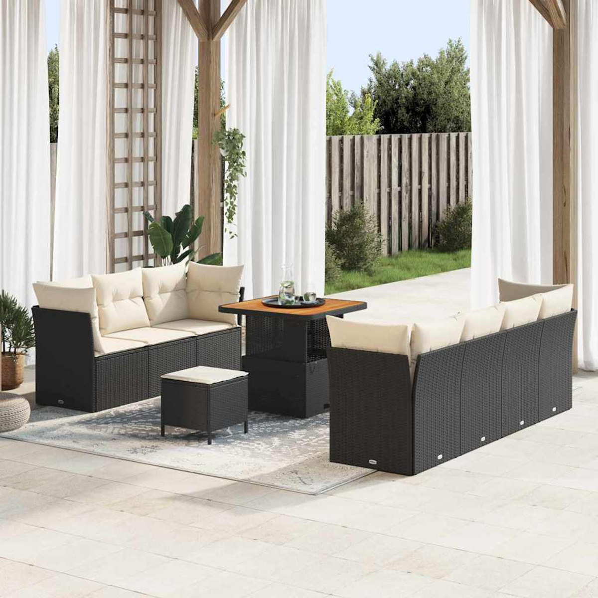 Set da patio: divani in vimini neri con cuscini beige, tavolo quadrato in legno e pouf su un tappeto, tende bianche sullo sfondo.