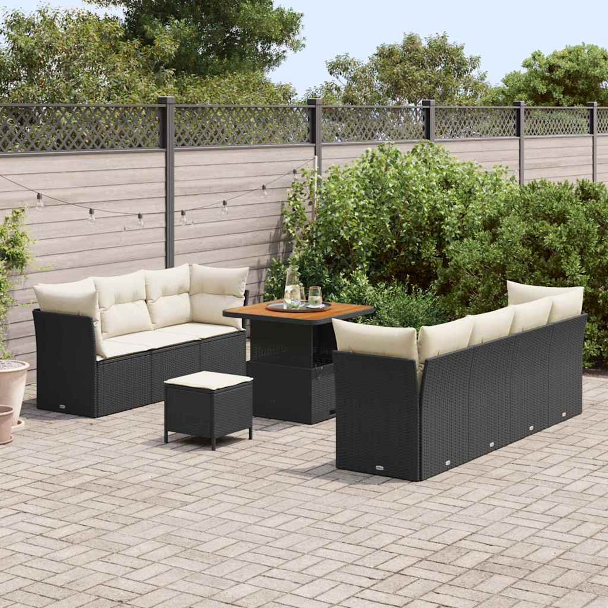 Set da patio esterno con mobili in vimini neri, cuscini beige e tavolo quadrato. Una recinzione in legno e verde sullo sfondo.