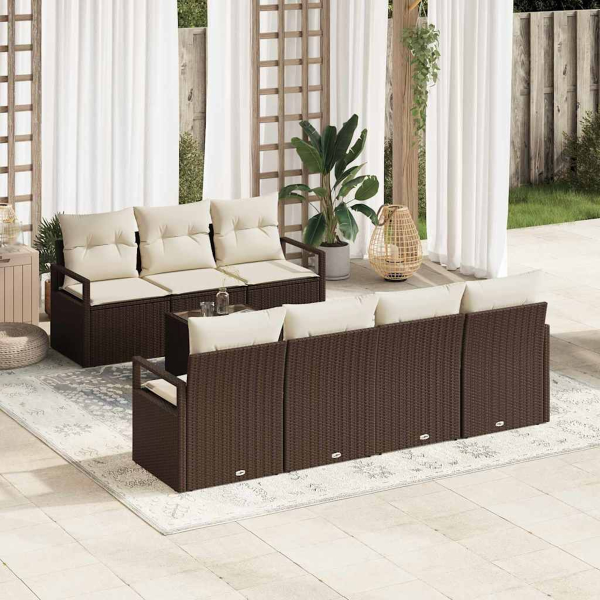 Juego de patio exterior con sofás de mimbre marrón, cojines crema y una alfombra. El fondo presenta cortinas blancas, plantas y un enrejado de madera.