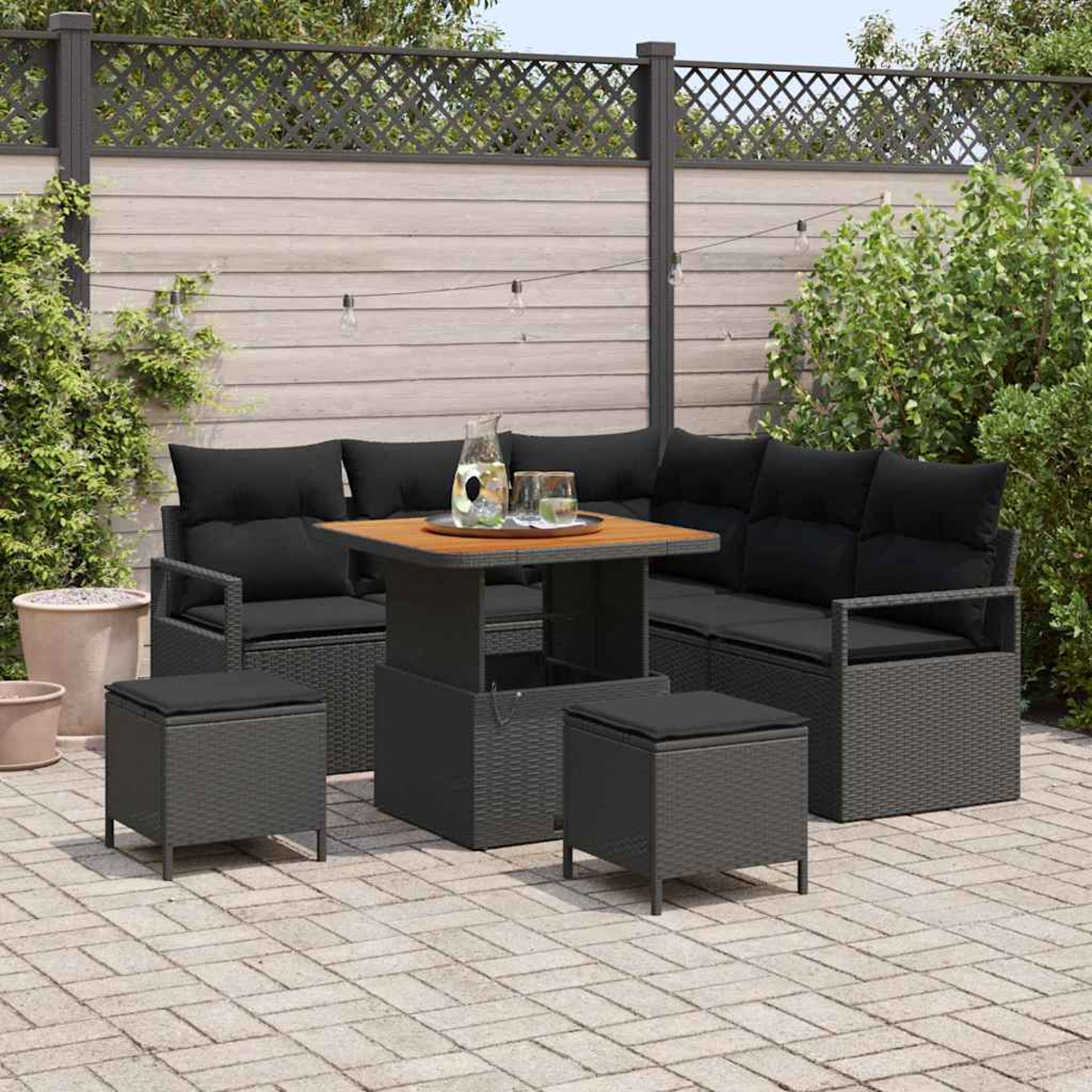Terrassenmöbel-Set im Freien mit schwarzen Kissen, einem Tisch und zwei Hockern auf einer Backstein-Patio. Holzzaun und Grün im Hintergrund.