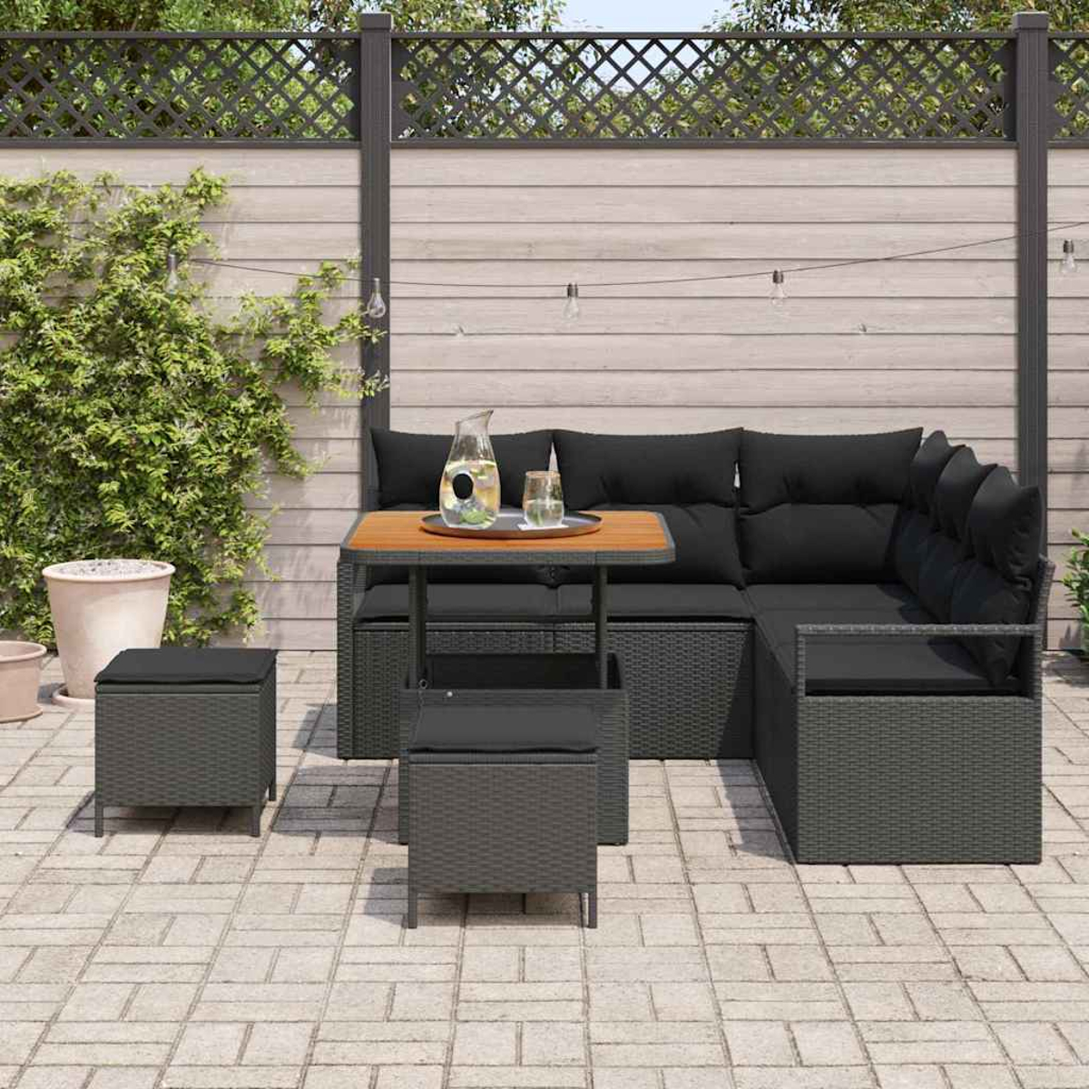 Garten-Terrassenset mit schwarzen Kissen, Tisch und Hockern. Auf dem Tisch stehen ein Krug und Gläser. Im Hintergrund ein Holzzaun.