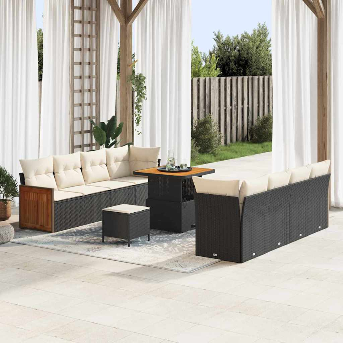 Außenbereich mit schwarzen Korbmöbeln, beigen Kissen und einem Holztisch unter weißen Vorhängen. Garten im Hintergrund.