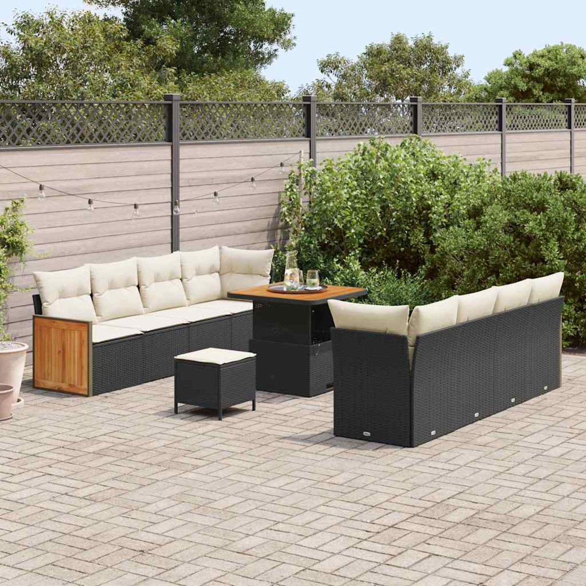 Gartenmöbel-Set mit schwarzen Korbs Sofas, einem Tisch und einem Hocker. Beige Kissen und ein Holzzaun sind im Hintergrund.