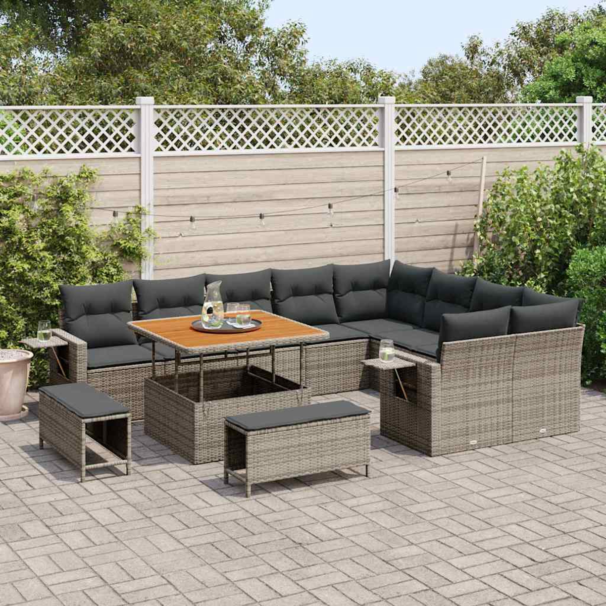 Ein Terrassenset mit großem Ecksofa, Tisch und zwei Bänken. Das Set ist grau mit schwarzen Kissen auf einer Backsteinterrasse mit Zaun.