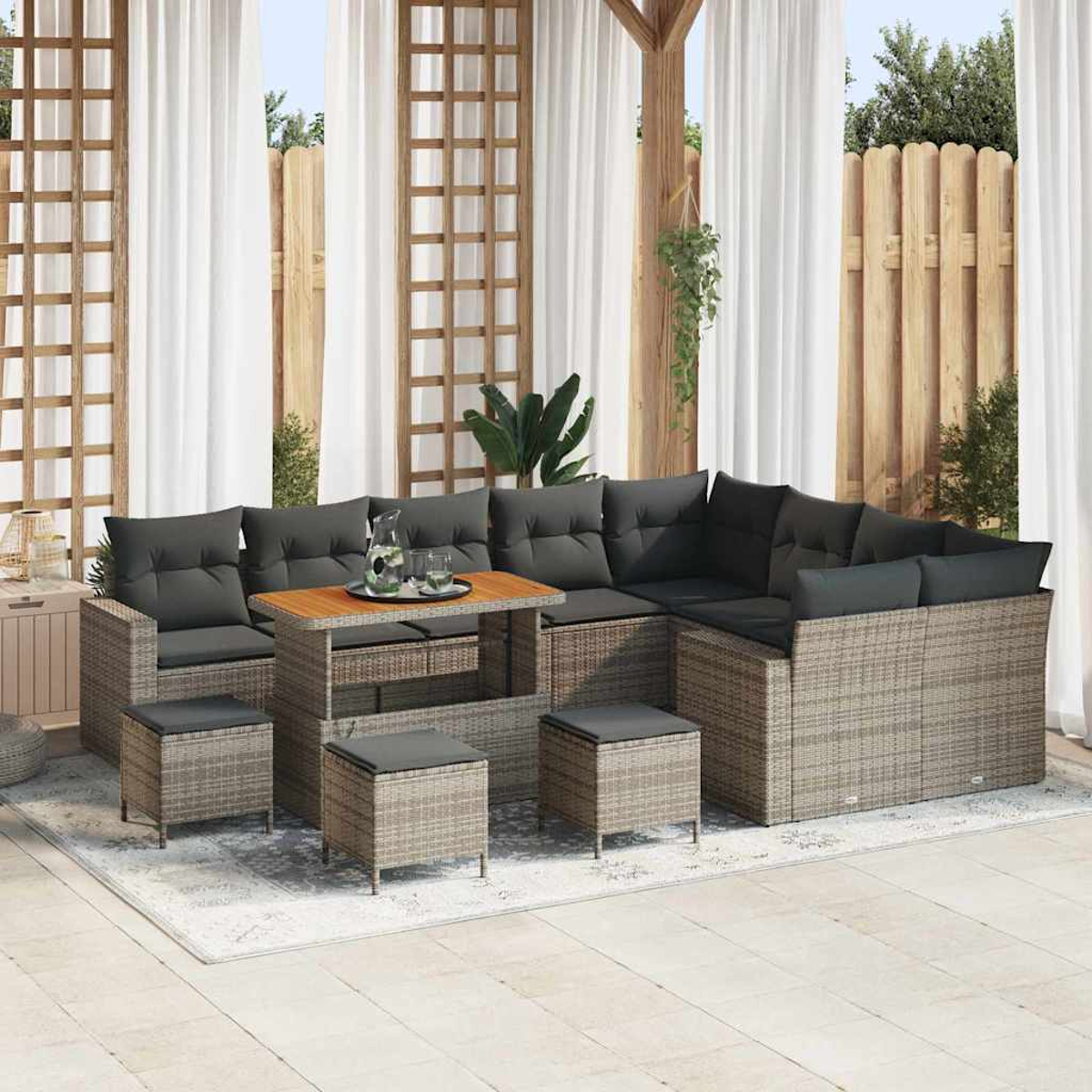 Garten-Sitzgruppe mit grauen Kissen, Holztisch und Hockern auf einem Teppich. Weiße Vorhänge und Holzzaun im Hintergrund.