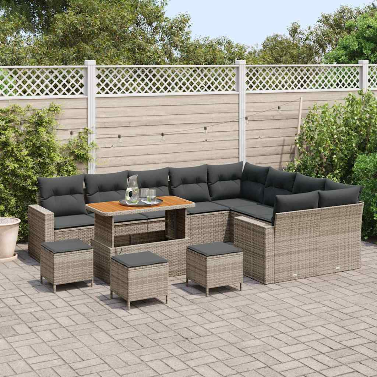 Outdoor-Ecksofa mit grauen Kissen, Holztisch mit Getränken und passenden Hockern auf einer Terrasse mit Zaun und Grün.