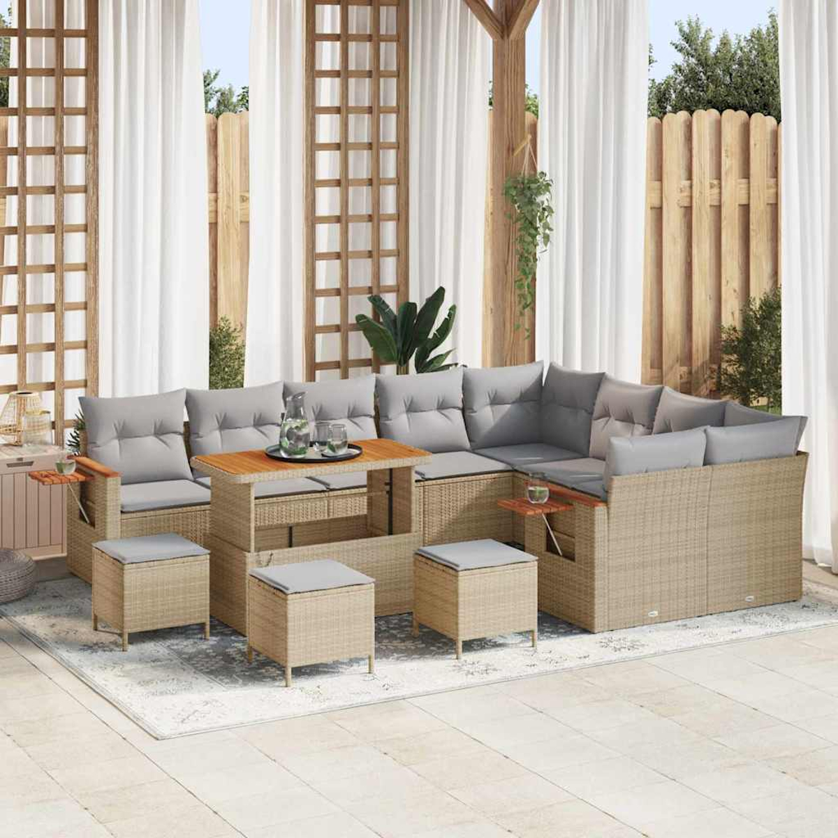 Sofá seccional para exteriores con cojines grises, otomanas a juego y una mesa de madera sobre una alfombra blanca. Cortinas blancas y valla de madera al fondo.