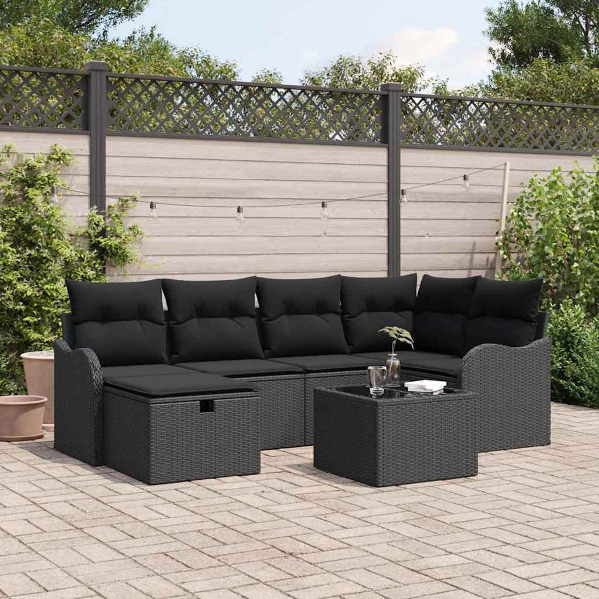 Schwarzes Outdoor-Korbsitzgarnitur mit Kissen, Tisch und Pflanzen auf einer Terrasse. Im Hintergrund ein Zaun und Grün.