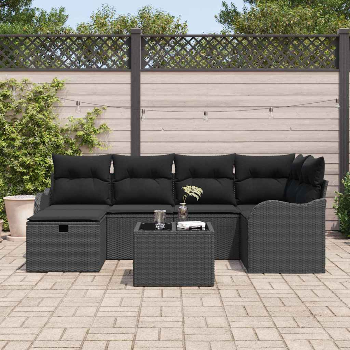Terrassenmöbel-Set mit grauem Korbmöbel, schwarzen Kissen und einem kleinen Tisch. Vor einem Holzzaun und Grün.