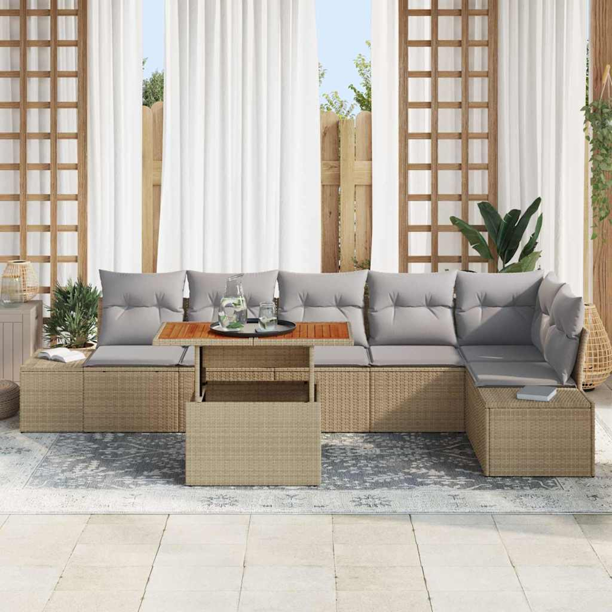 Zona de estar al aire libre con muebles de mimbre beige, cojines grises, mesa de madera y cortinas blancas. Plantas y una alfombra completan la escena.