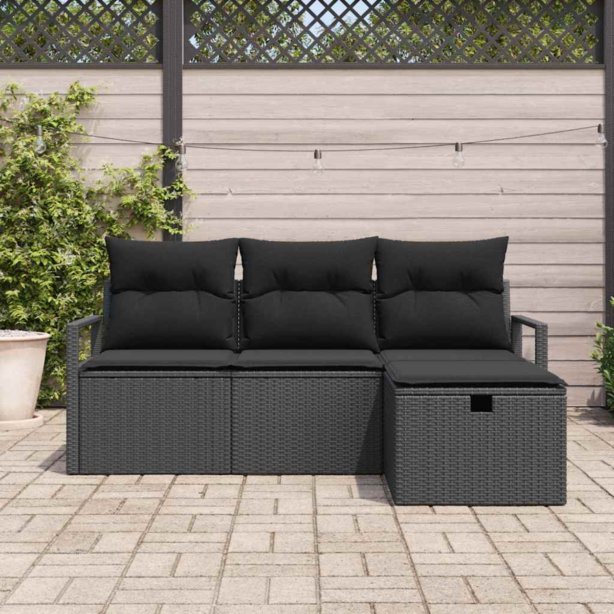 Ein dunkelgraues Outdoor-Ecksofa aus Korbgeflecht mit schwarzen Kissen steht auf einer Ziegelterrasse. Dahinter ein Spalier und ein Holzzaun.