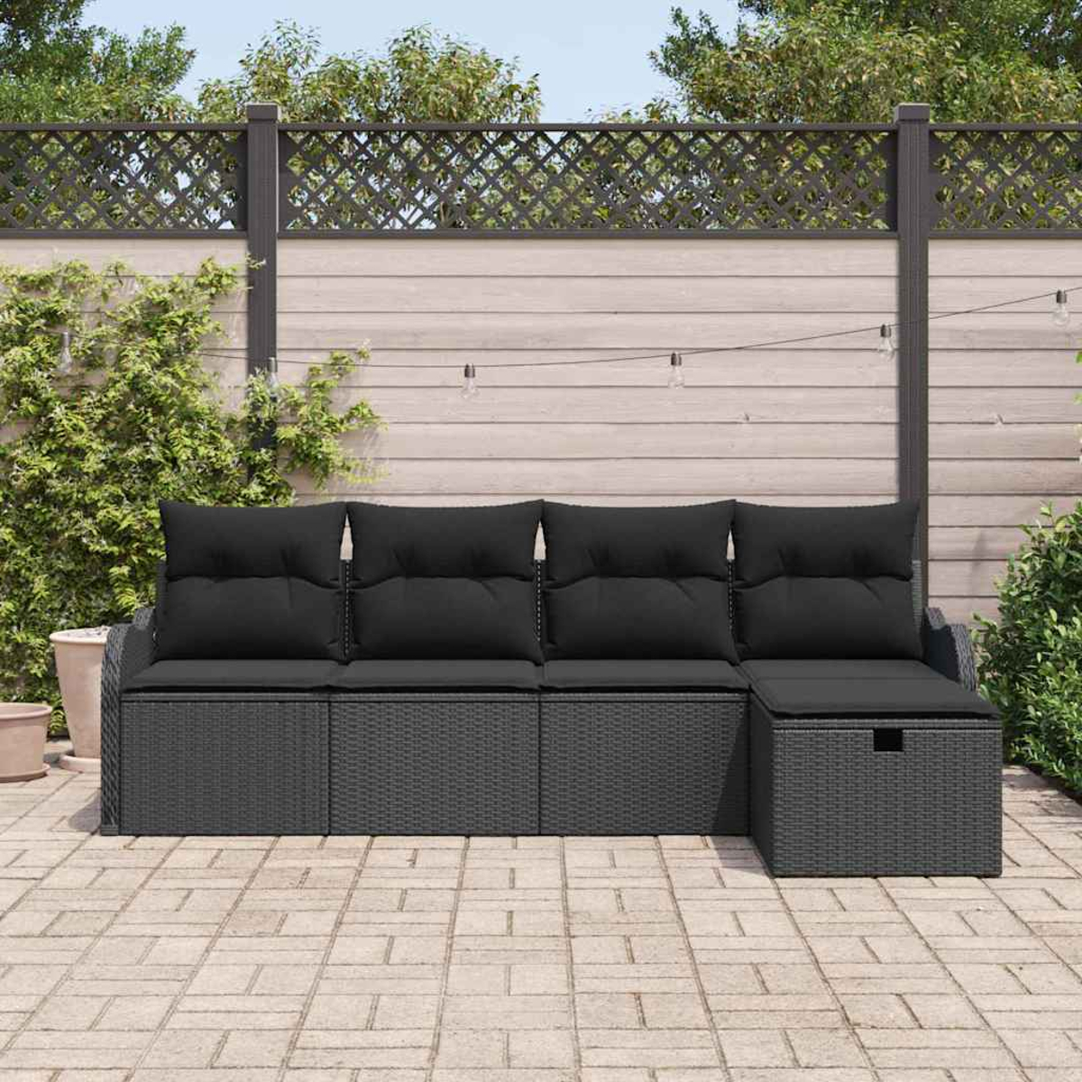 Ein graues Outdoor-Sofa aus Korbgeflecht mit schwarzen Kissen steht auf einer Backstein-Terrasse. Ein Holzzaun und Grünzeug sind im Hintergrund.