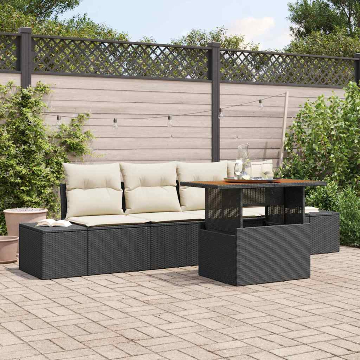 Garten-Terrassenset mit schwarzen Korbmöbeln, beigefarbenen Kissen und einem Holztisch. Im Hintergrund ein Zaun und Grün.