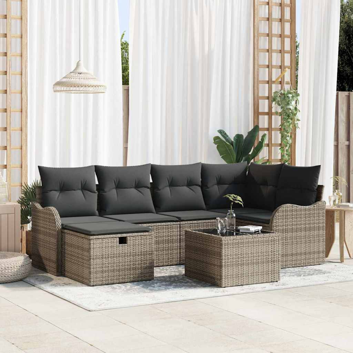 Gartenmöbel-Set aus Korbgeflecht mit grauen Kissen. Enthält ein Sofa, eine Ottomane und einen kleinen Tisch. Weiße Vorhänge und Pflanzen im Hintergrund.