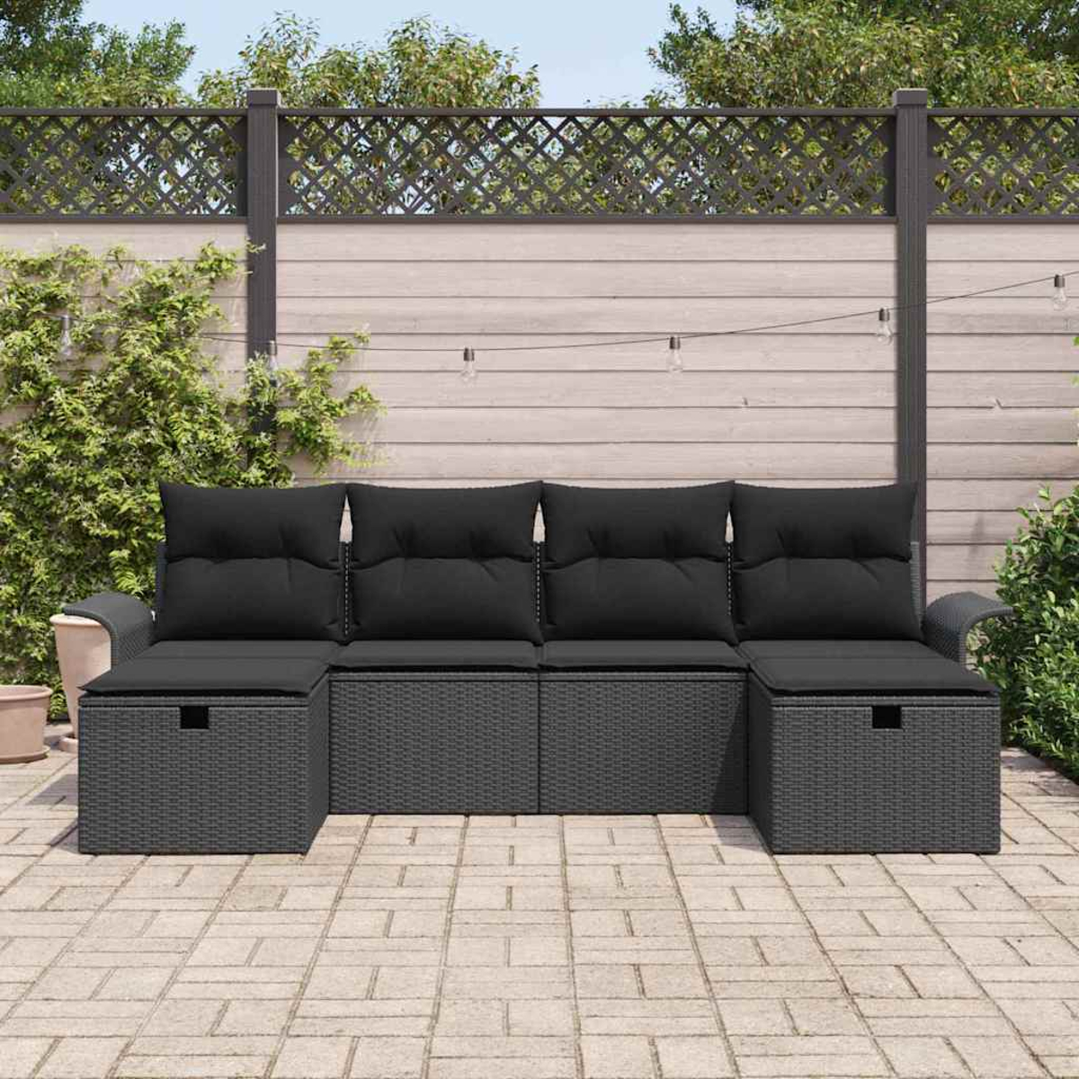 Outdoor-Ecksofa mit schwarzen Kissen. Graue Korbbasis. Hintergrund hat Holzzaun, Grün und Lichterketten.
