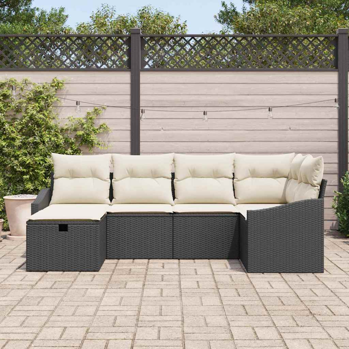 Outdoor-Ecksofa mit cremefarbenen Kissen. Dunkelgrauer Korbrahmen. Dahinter ein Holzzaun und Lichterketten. Steinterrasse.