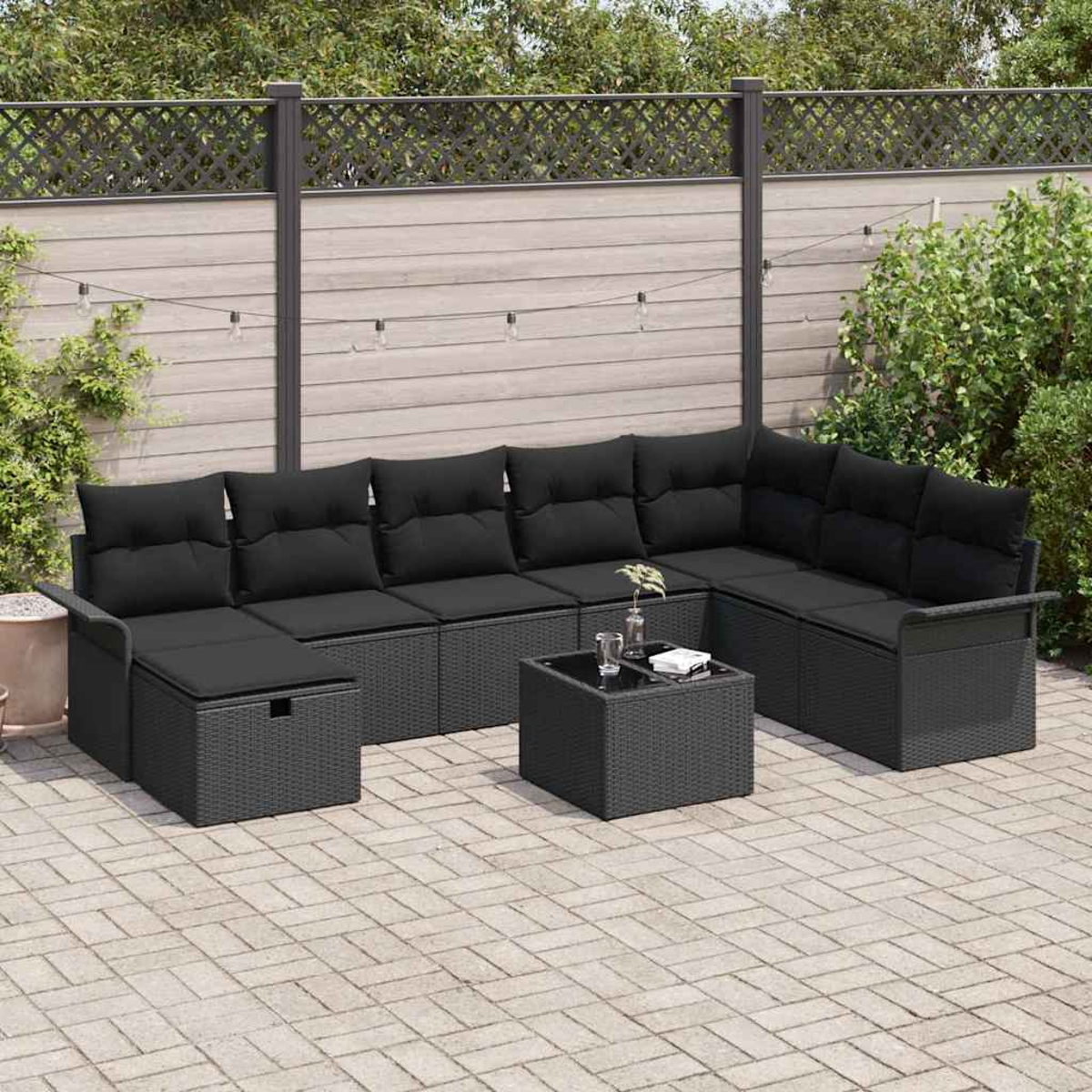 Ein Ecksofa im Freien mit schwarzen Kissen und einem passenden Couchtisch steht auf einer Ziegelterrasse. Im Hintergrund befinden sich ein Zaun und Grün.
