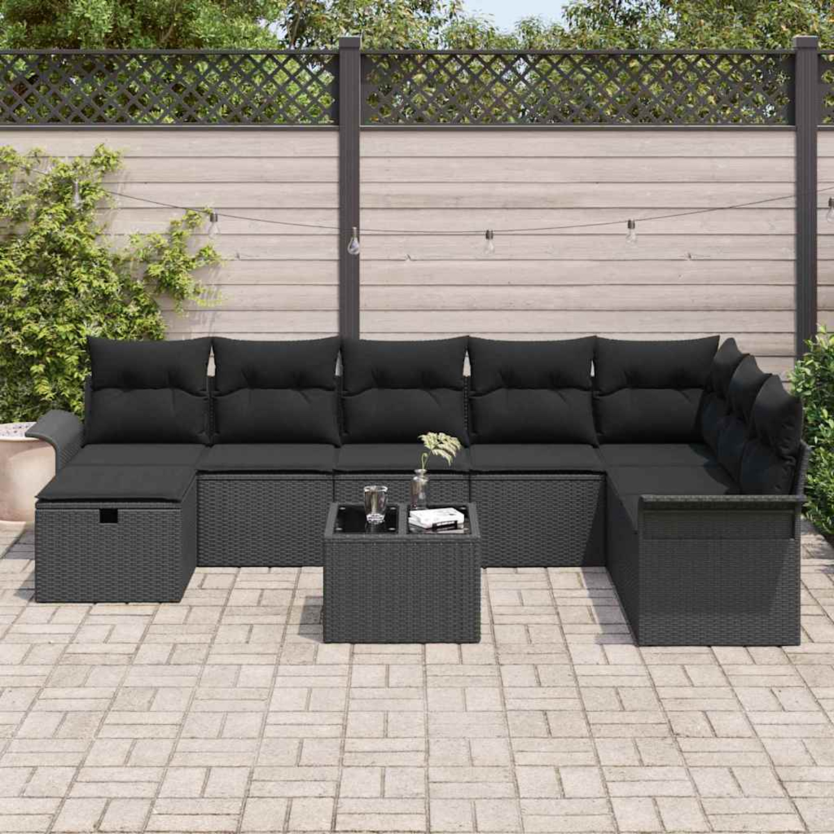 Outdoor-Ecksofa mit schwarzen Kissen, einem kleinen Tisch und einer Ziegelterrasse. Im Hintergrund ein Holzzaun und Laub.