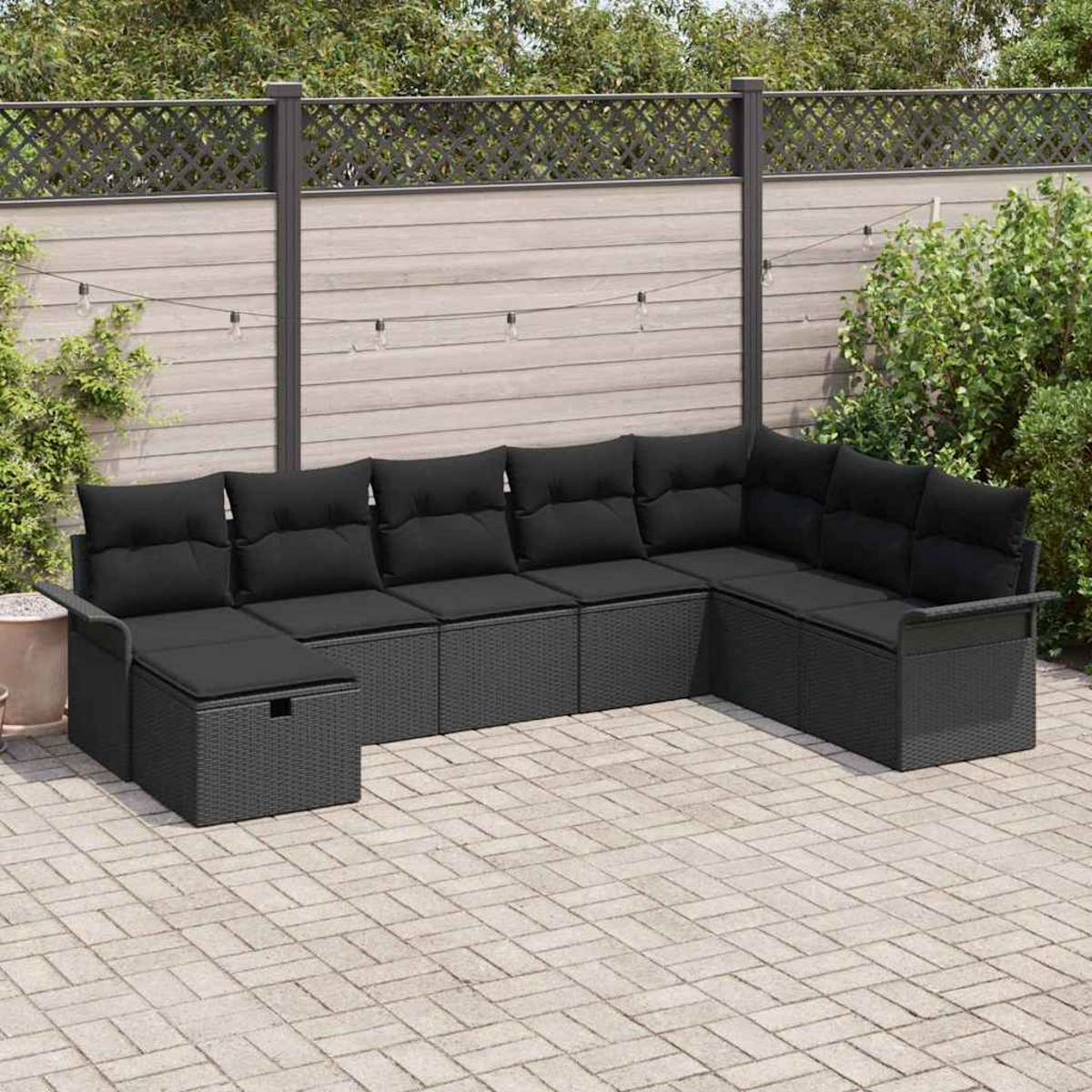 Ein graues Ecksofa mit schwarzen Kissen steht auf einer Backsteinterrasse. Im Hintergrund befinden sich ein Zaun und Grün.