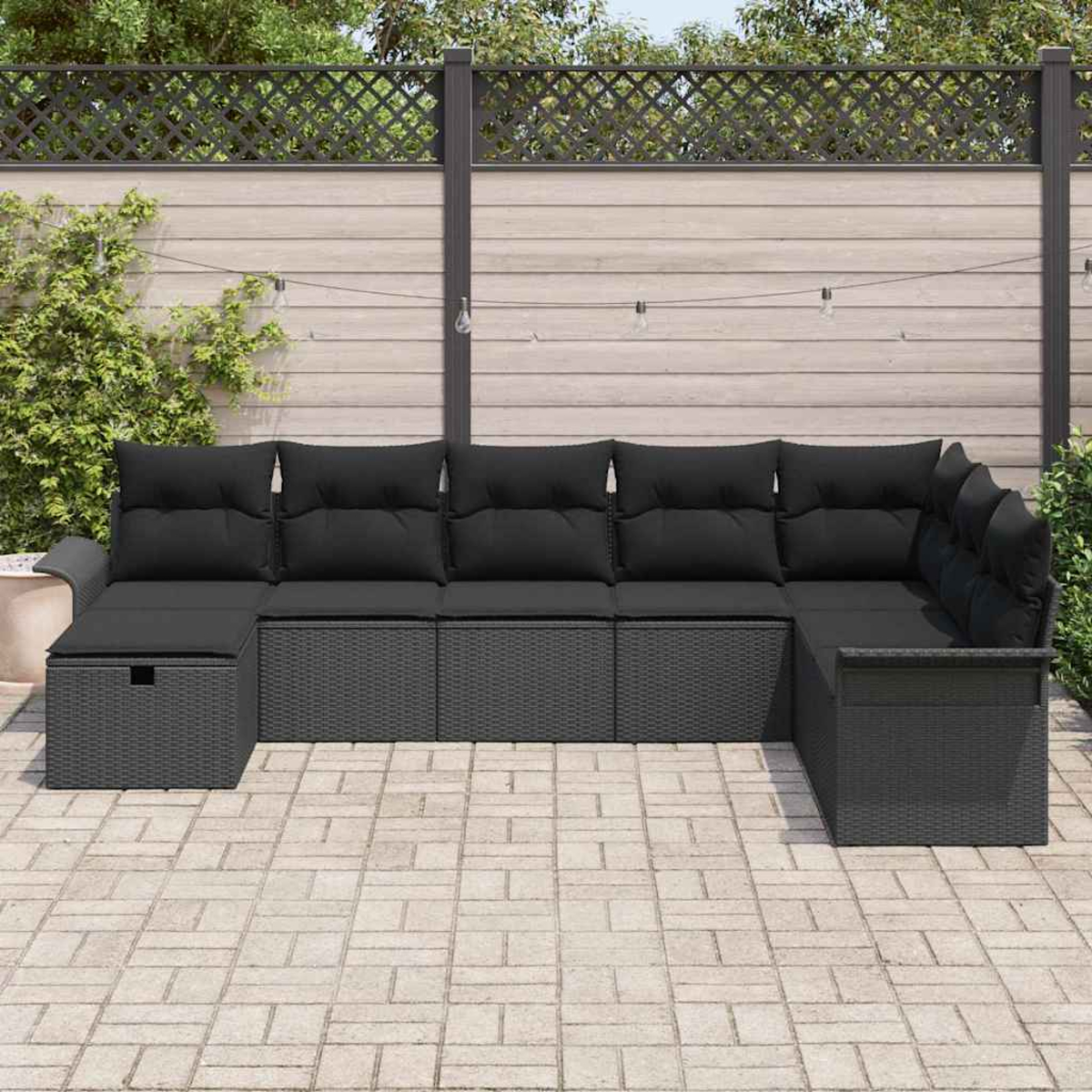 Ein Ecksofa im Freien mit schwarzen Kissen. Das Sofa ist dunkelgrau und steht auf einer Ziegelterrasse mit Holzzaun im Hintergrund.