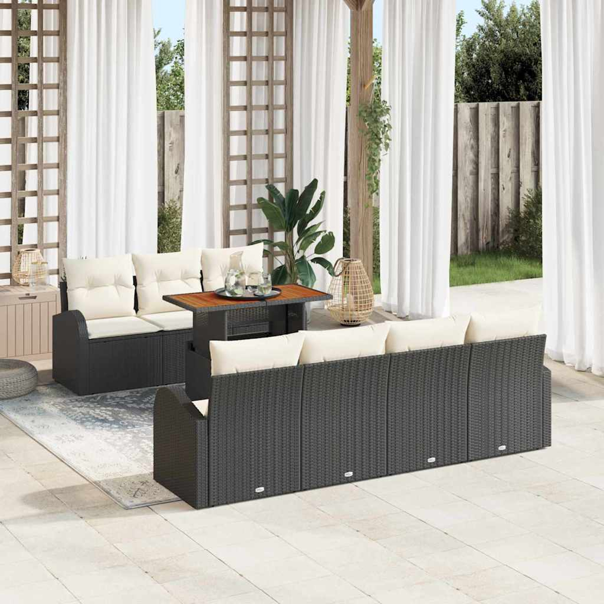Terrasse mit dunkelgrauen Korbmöbeln, cremefarbenen Kissen, Holztisch und weißen Vorhängen. Hintergrund mit Grün und Holzzaun.