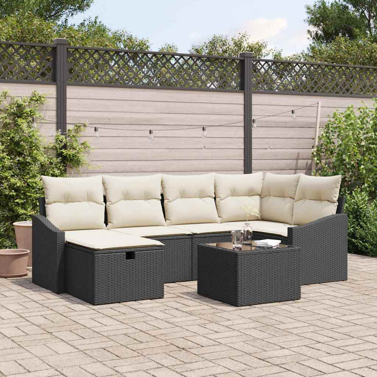 Outdoor-Ecksofa mit beigen Kissen. Davor steht ein kleiner quadratischer Tisch. Im Hintergrund ein Zaun und Laub.