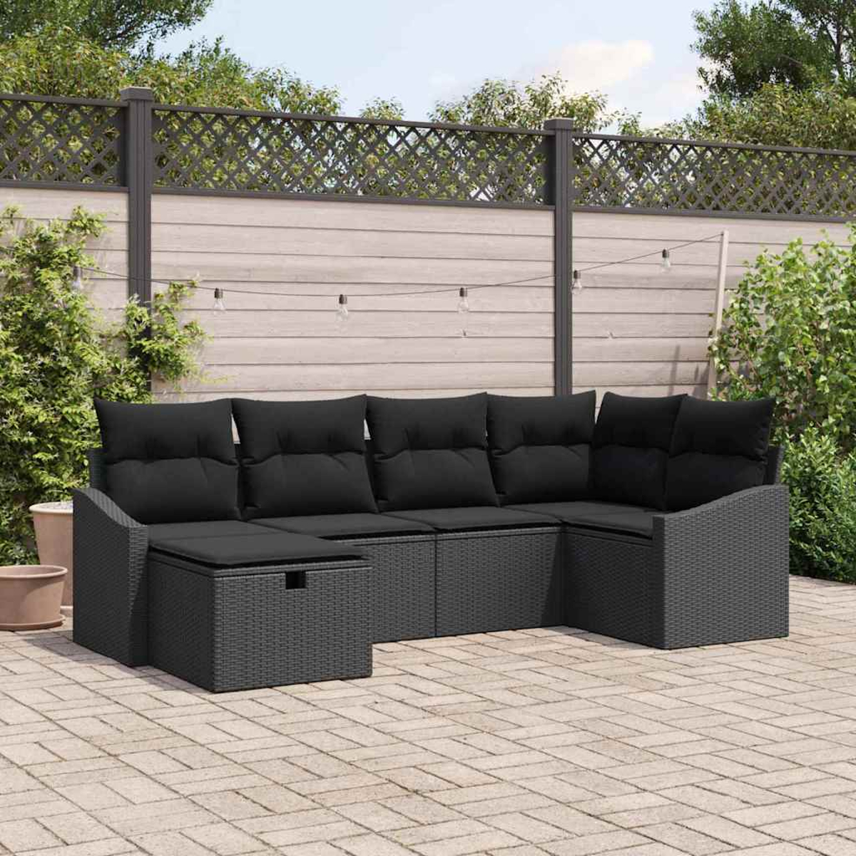 Schwarzes Outdoor-Wicker-Ecksofa mit schwarzen Kissen auf einer Backstein-Terrasse. Im Hintergrund ein Zaun mit Lichtern und Pflanzen.