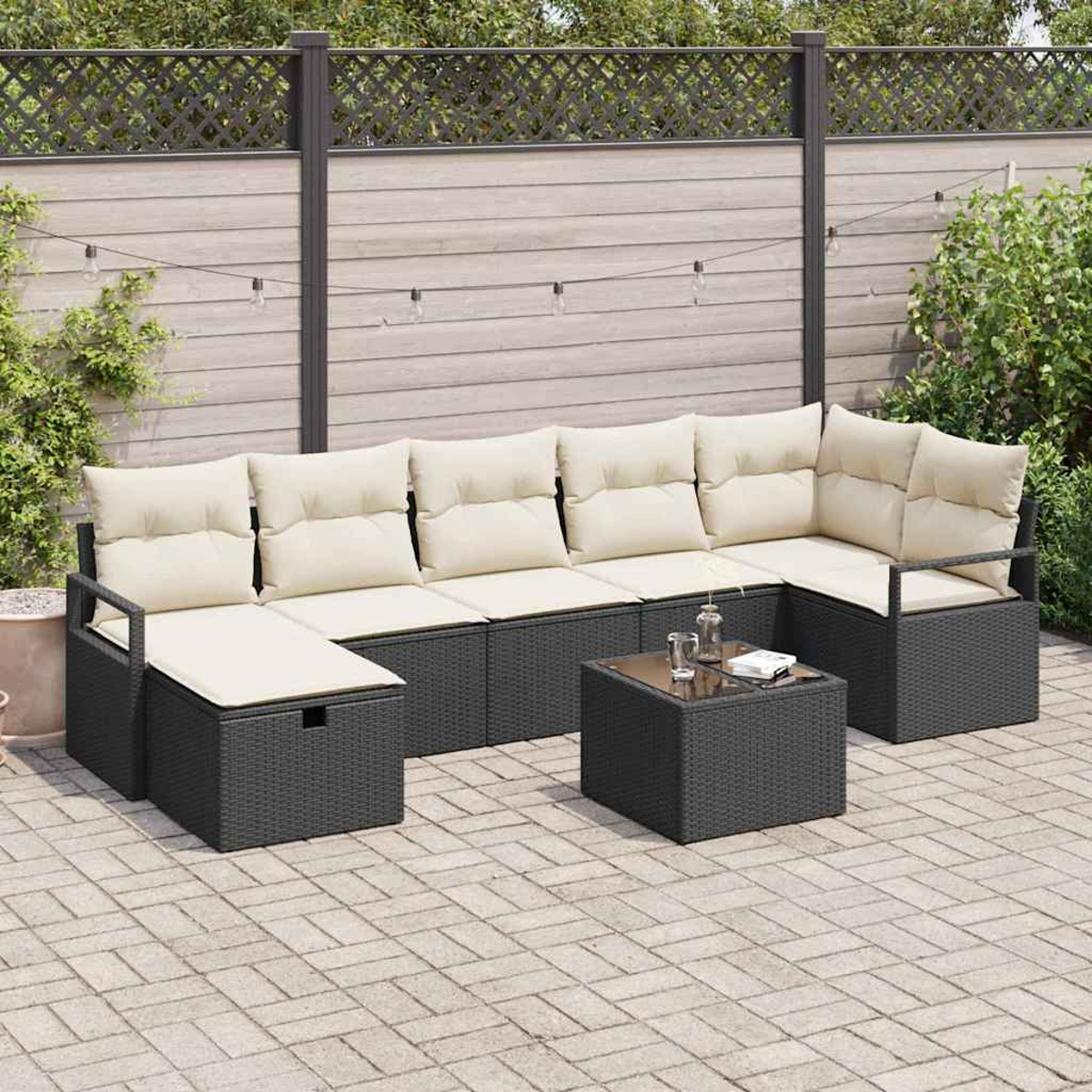 Outdoor-Ecksofa mit cremefarbenen Kissen, schwarzem Korbrahmen und Tisch mit Glasplatte auf einer Backstein-Terrasse, mit Zaun im Hintergrund.