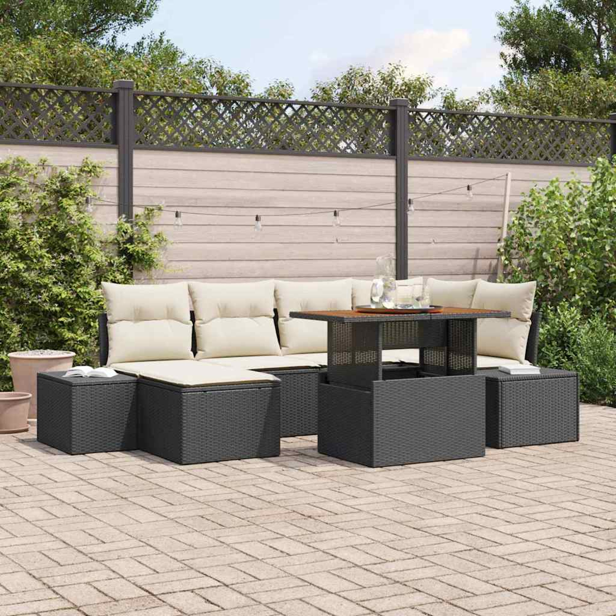Garten-Terrassenset: dunkelgraues Korbmöbel-Sofa, Ottomane und Tisch mit cremefarbenen Kissen. Holzzaun mit Lichterkette im Hintergrund.