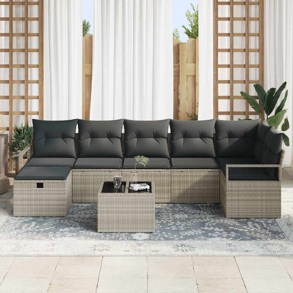 Outdoor-Sitzgruppe mit grauen Kissen, einem kleinen Tisch und einem gemusterten Teppich. Weiße Vorhänge und Holzgitter im Hintergrund.
