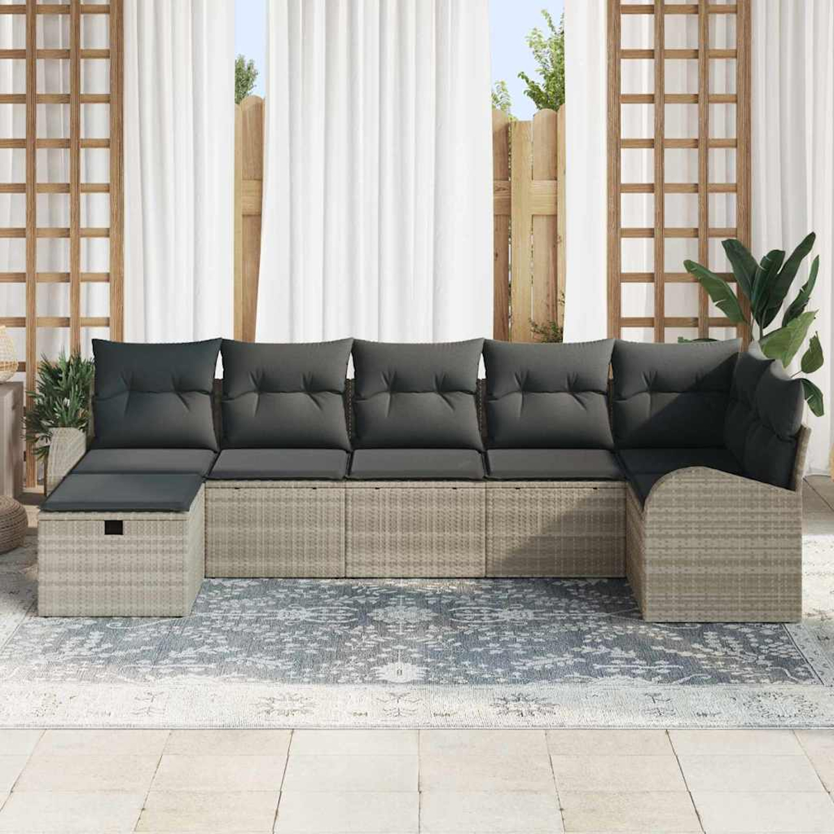 Ein Ecksofa im Freien mit grauen Kissen auf einem gemusterten Teppich, vor weißen Vorhängen und einem Holzgitter.