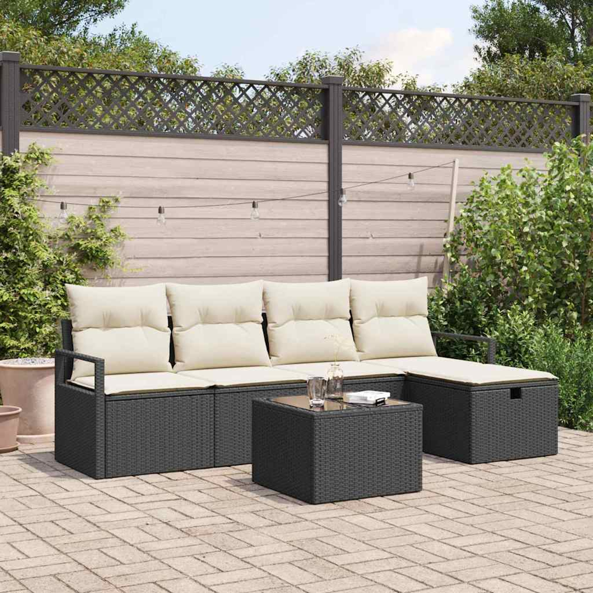 Garten-Set mit dunkelgrauen Korbmöbeln, beigen Kissen und einem Tisch mit Glasplatte. Vor Holzzaun und Grün.