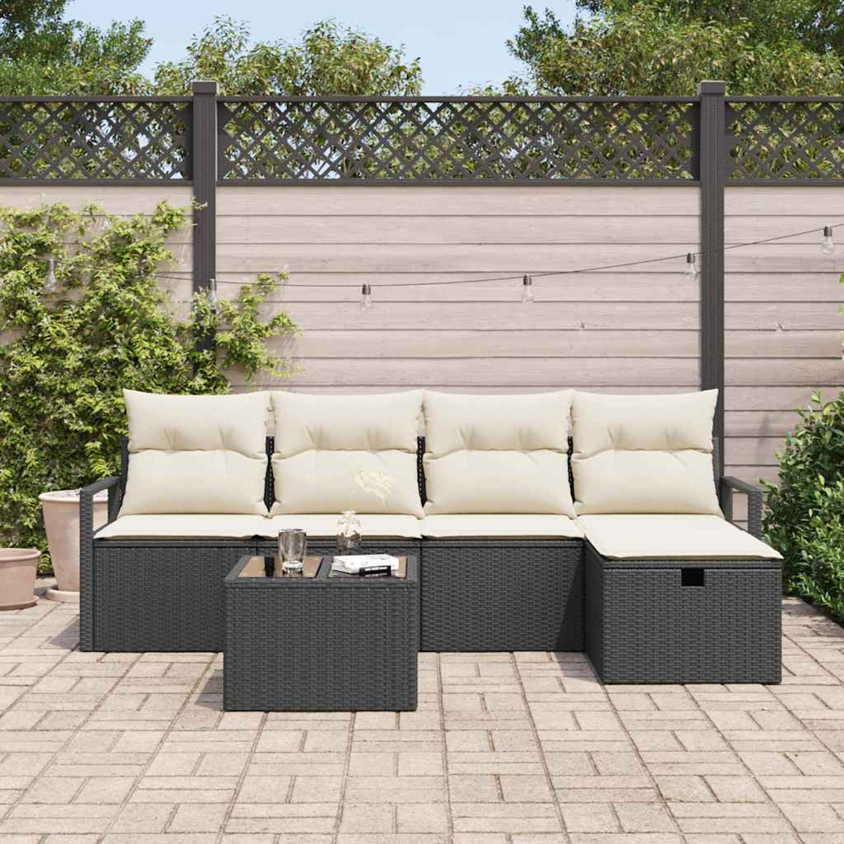 Garten-Sitzgruppe mit grauem Korbgeflecht, cremefarbenen Kissen und einem passenden Tisch auf einer Ziegelterrasse, im Hintergrund ein Holzzaun.