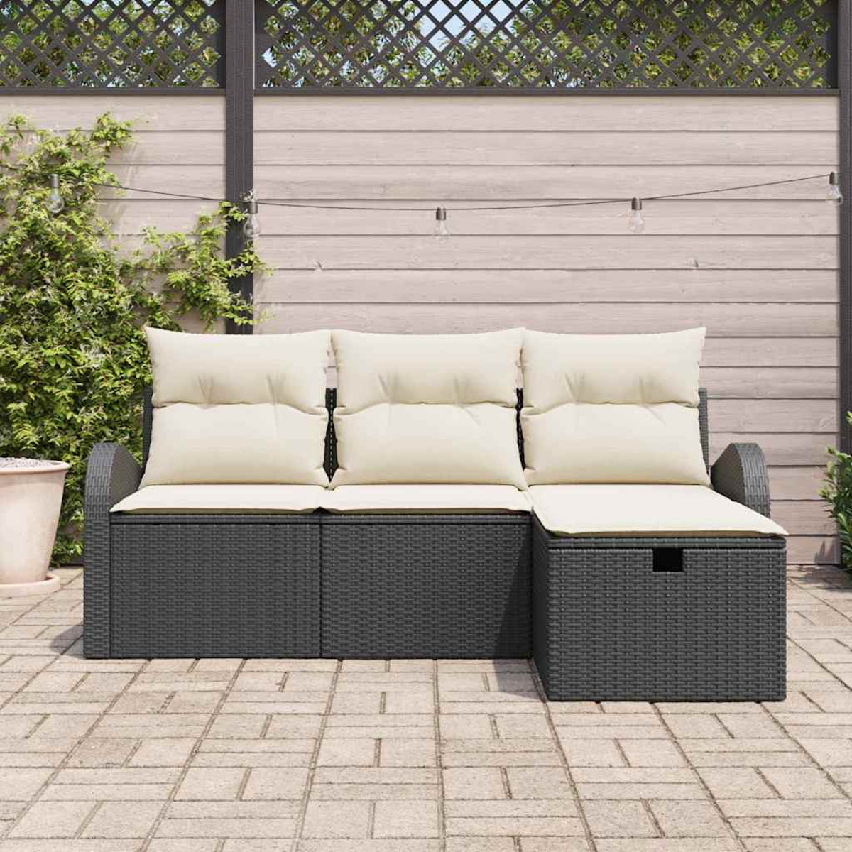 Dunkelgraues Outdoor-Sofa aus Korbgeflecht mit cremefarbenen Kissen. Holzzaun und Lichterketten im Hintergrund. Gepflasterte Terrasse.