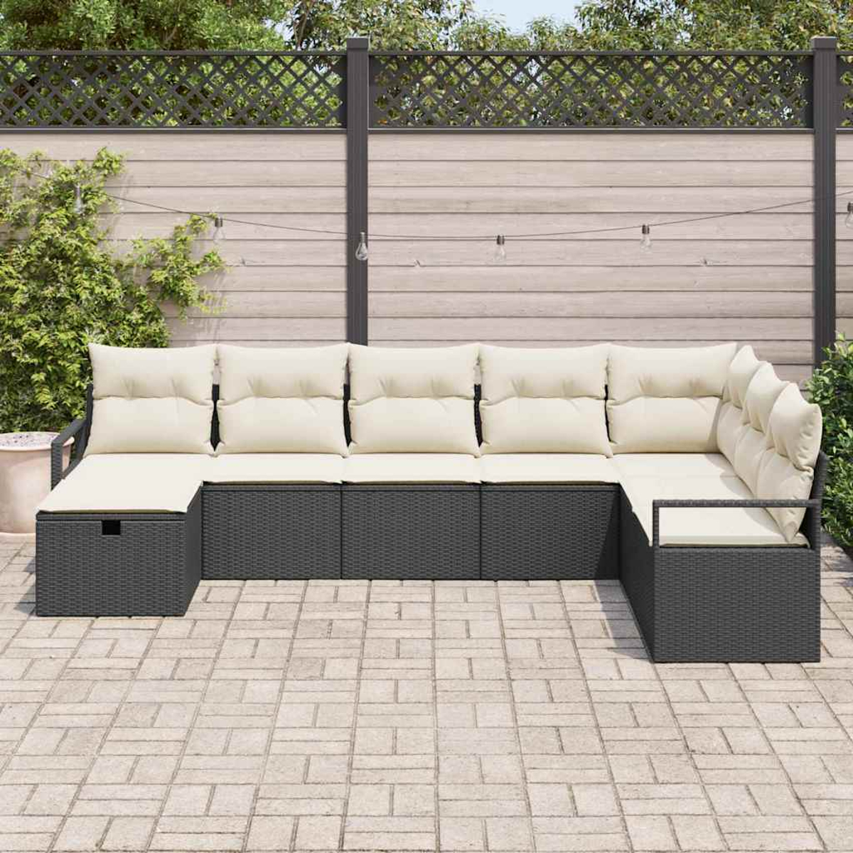 Dunkelgraues Outdoor-Ecksofa mit cremefarbenen Kissen steht auf einer Backstein-Terrasse. Im Hintergrund ein Holzzaun und ein Spalier.