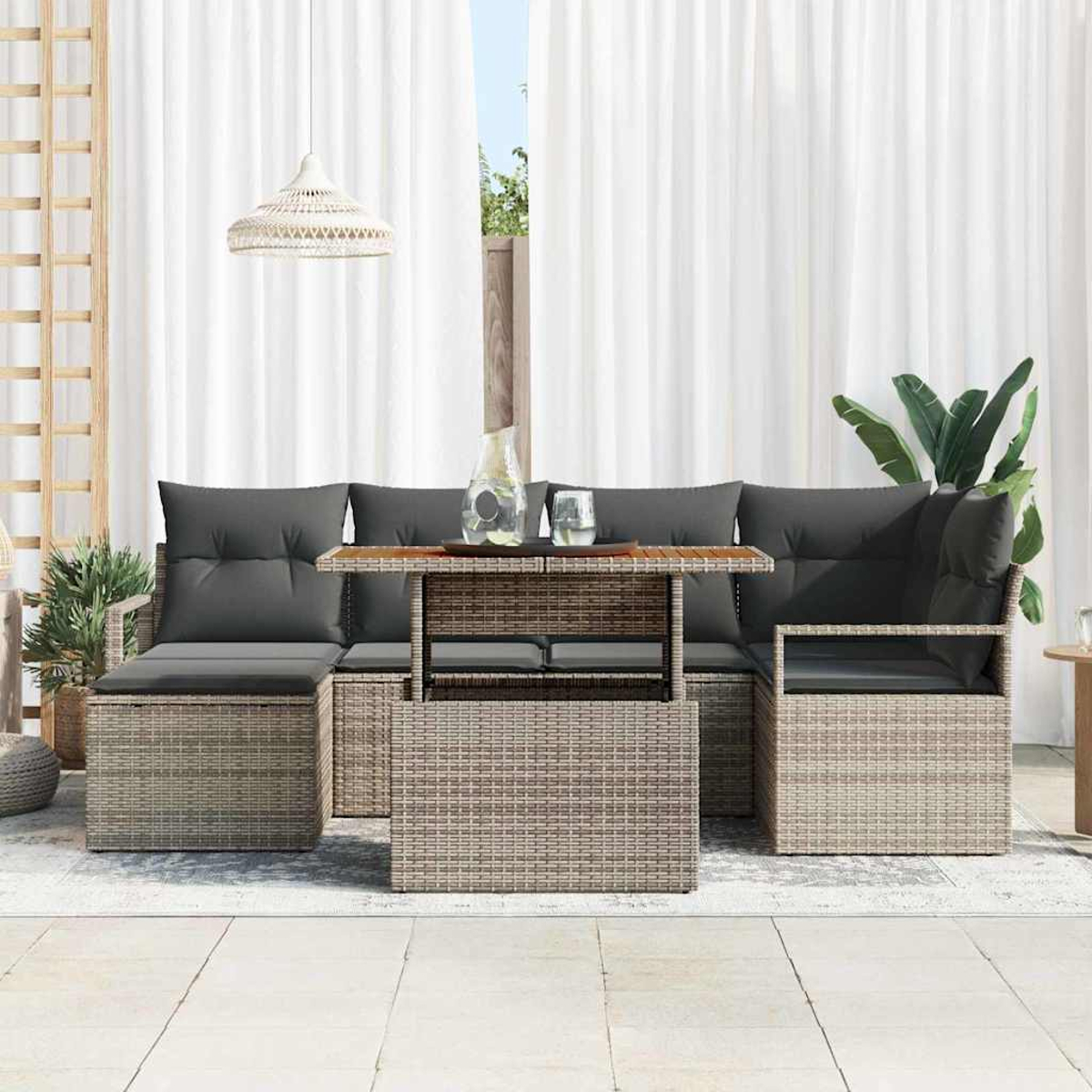Juego de muebles de mimbre para exteriores con cojines grises. Incluye un sofá, una mesa y una silla. Cortinas blancas y plantas en el fondo.
