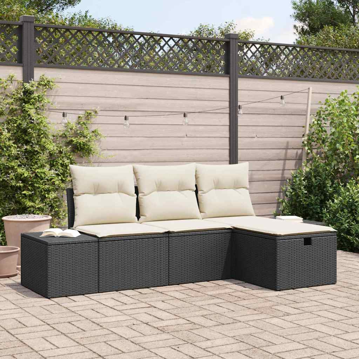 Ein schwarzes Rattan-Ecksofa mit cremefarbenen Kissen steht auf einer Ziegelterrasse. Ein Holzzaun und Grün bilden den Hintergrund.
