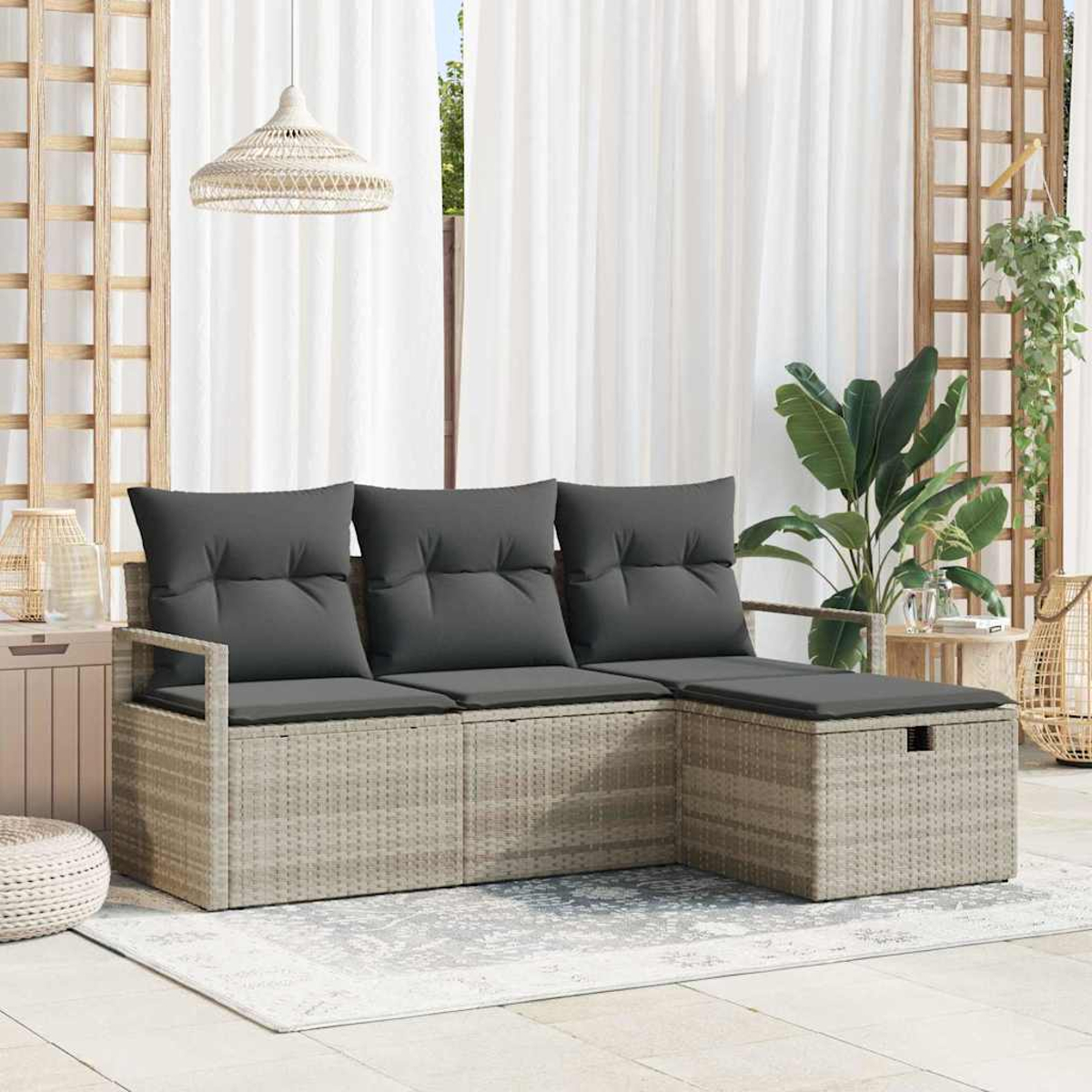 Sofá seccional de mimbre con cojines grises. Fondo: cortinas blancas, plantas y una lámpara colgante. Alfombra en el suelo.
