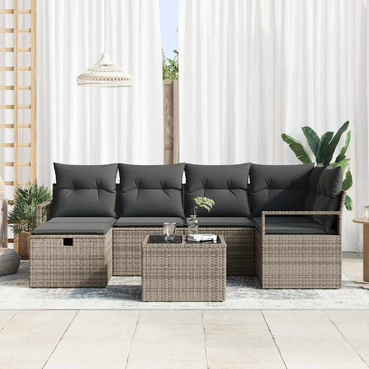 Gartensofa mit grauen Kissen und einem kleinen Tisch auf einer Terrasse. Weiße Vorhänge und Pflanzen im Hintergrund.