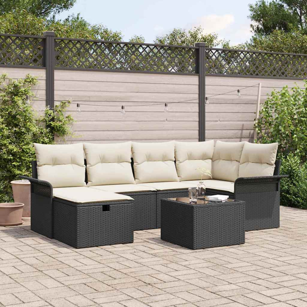 Schwarzes Outdoor-Wicker-Sofaset mit beigen Kissen und einem kleinen Tisch auf einer Terrasse. Hintergrund: Zaun und Pflanzen.