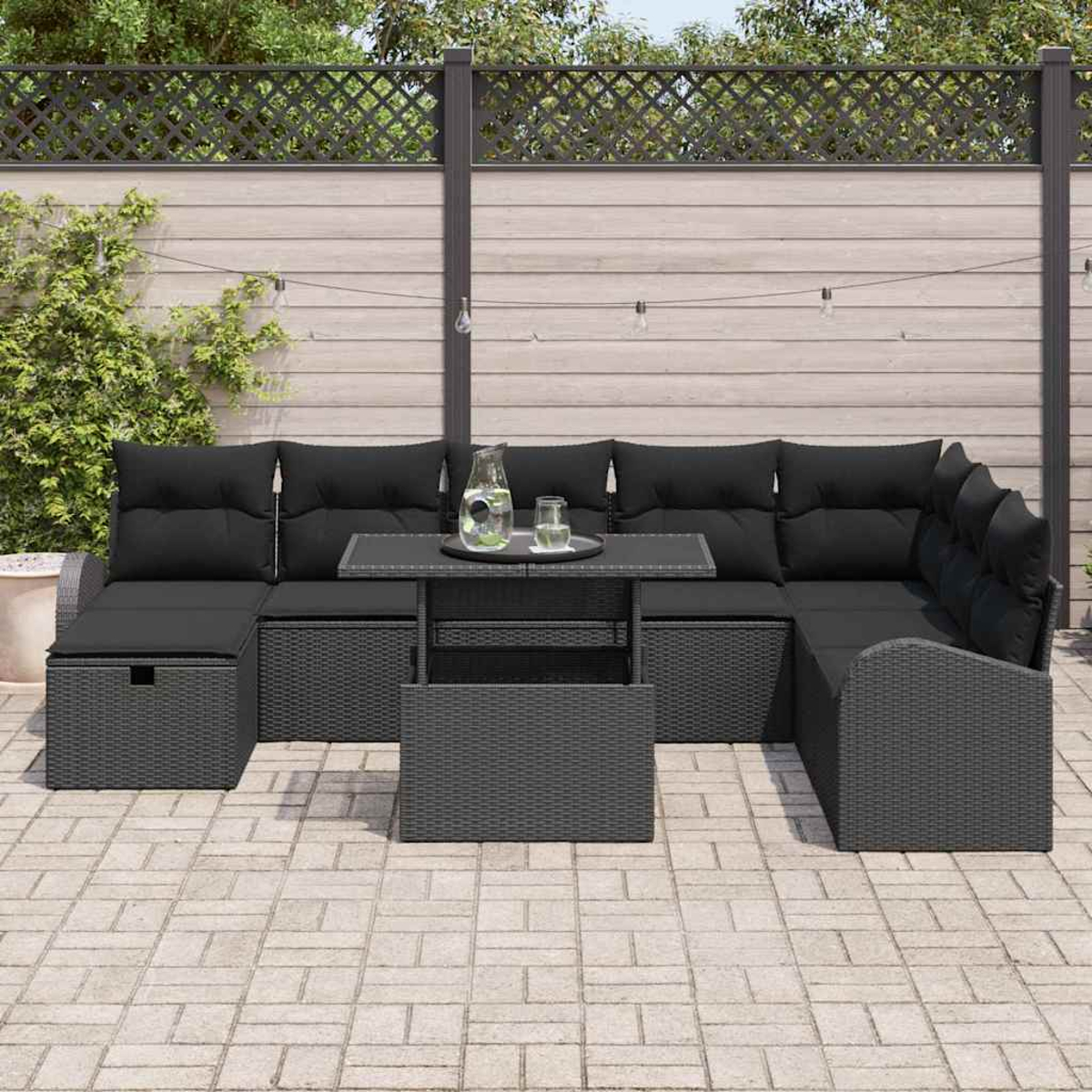 Ein schwarzes Outdoor-Sofa aus Korbgeflecht mit Kissen, ein passender Tisch mit Tablett auf einer gepflasterten Terrasse. Im Hintergrund ein Zaun.