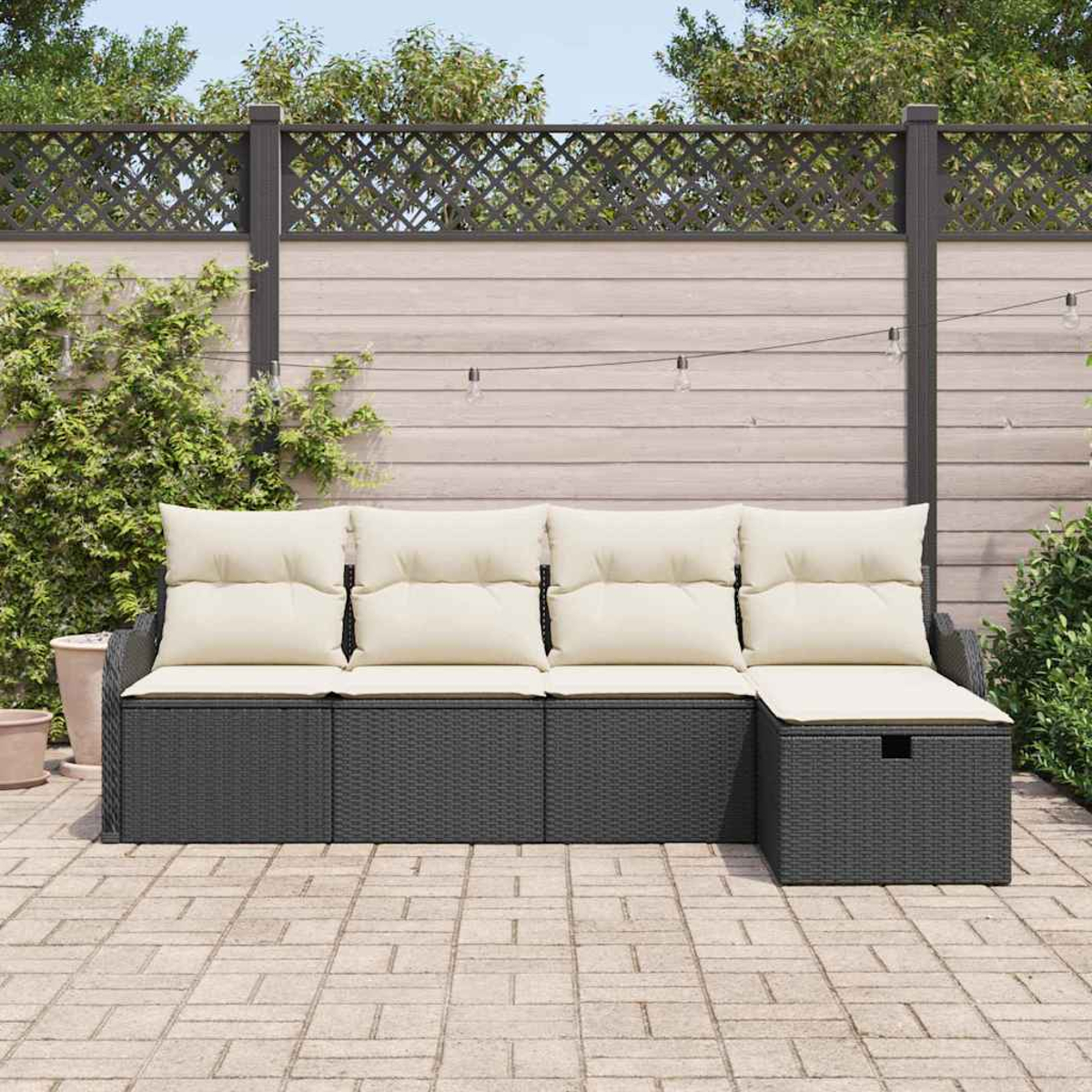 Gartensektionssofa mit cremefarbenen Kissen auf grauem Untergestell. Ziegelterrasse davor, Holzzaun und Grün im Hintergrund.