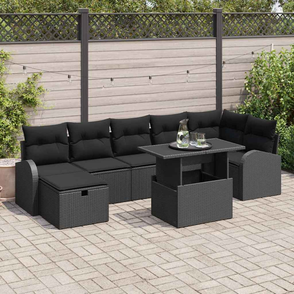 Terrassenset mit schwarzen Kissen, Tisch und Krug mit Gläsern. Auf einer Ziegelterrasse, Zaun im Hintergrund.