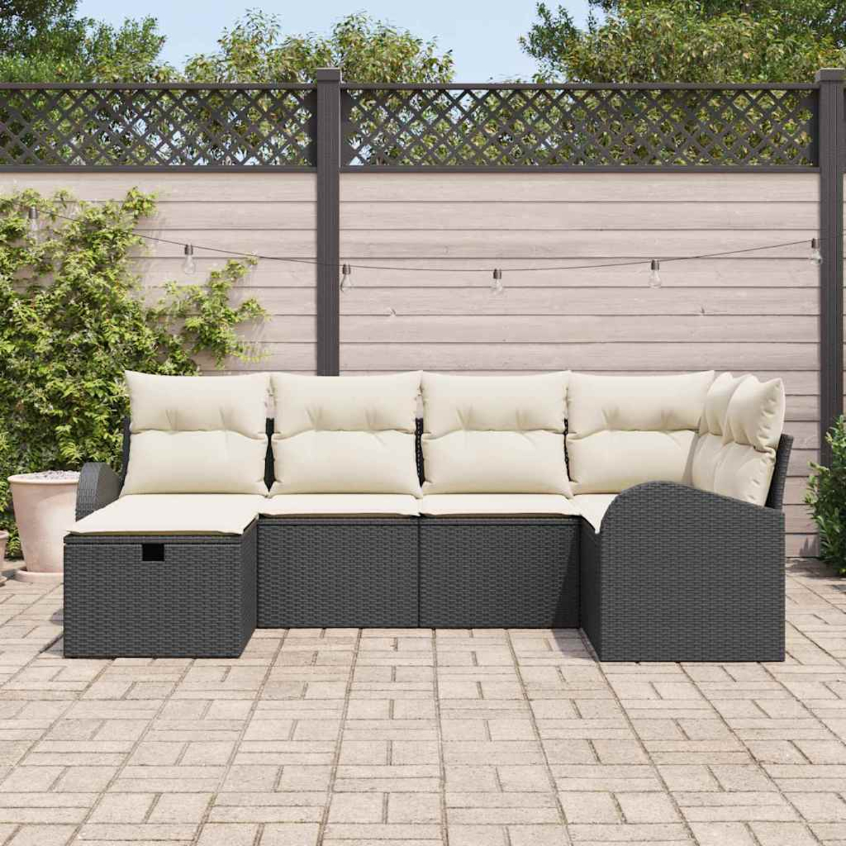 Outdoor-Ecksofa mit cremefarbenen Kissen. Grauer Korbrahmen. Terrasse mit Holzzaun und Grün im Hintergrund.
