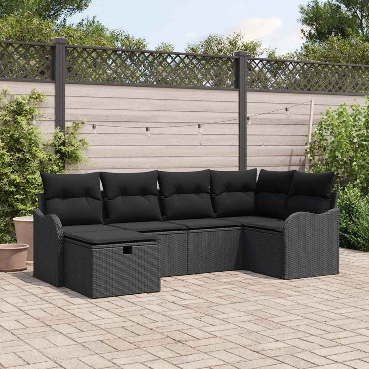Ein schwarzes Outdoor-Ecksofa aus Korbgeflecht mit schwarzen Kissen steht auf einer Backstein-Terrasse. Im Hintergrund befinden sich ein Zaun und Grün.